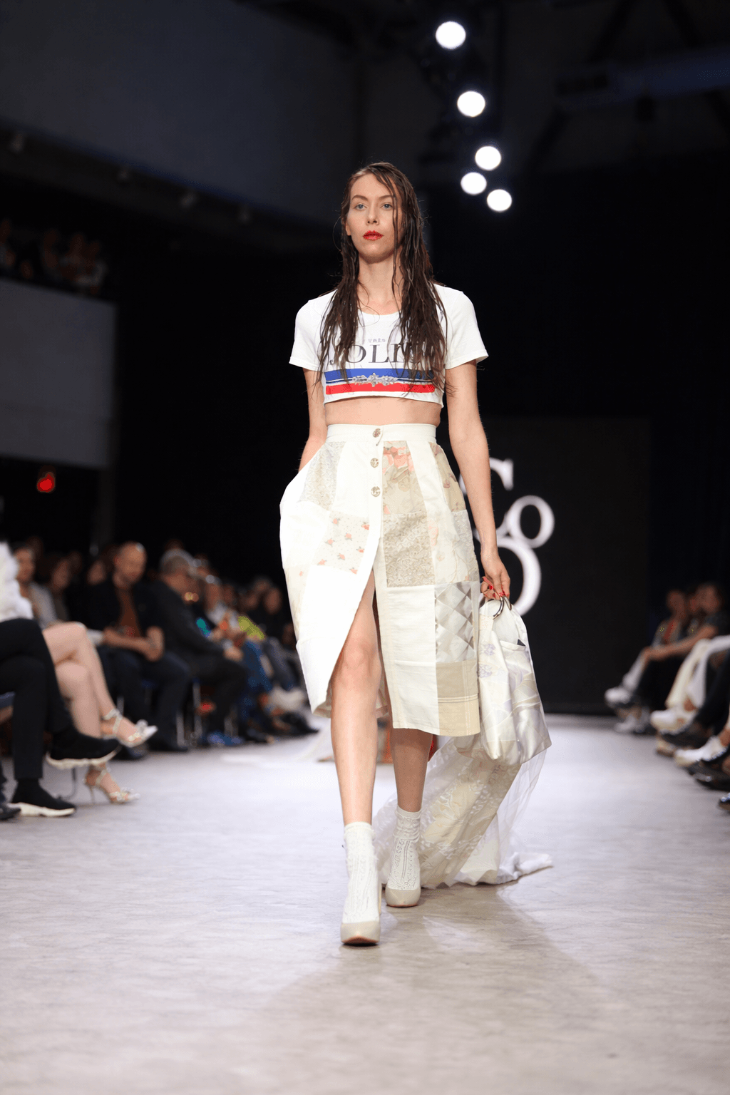 Model leading the runway in a white graphic crop top and a patchwork skirt trailing a long white fabric train